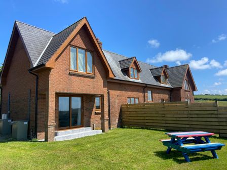 A brick house with multiple windows and a wooden fence on a grassy lawn with a colorful picnic table at Northcott View in Poughill near Bude
