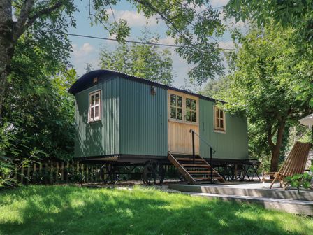 A green shepherd hut on wheels with wooden doors and steps in a garden with trees and lawn at The Happy Hut in Sopley near Christchurch