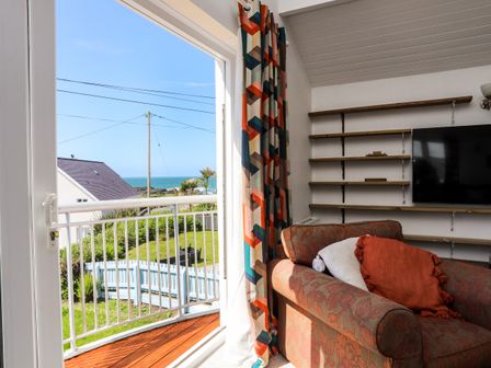 A room with a patterned armchair and cushions next to a glass door leading to a balcony overlooking a garden and the sea at Seaside Sanctuary in Trearddur Bay