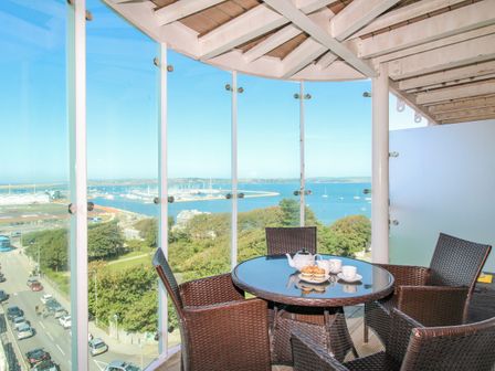 A balcony with a round glass table and wicker chairs overlooking a marina and trees at Sunset View in Castletown Portland