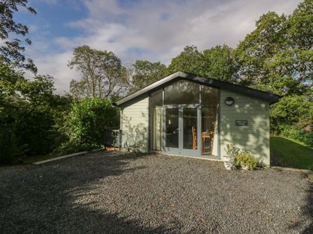 A small lodge with glass doors and chairs outside surrounded by trees at Bay Tree Lodge in Cartmel