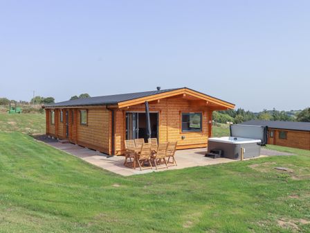 A wooden lodge with patio furniture and a hot tub on a grassy lawn at Foxglove Lodge in Hittisleigh