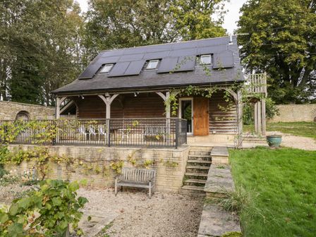 A wooden house with solar panels on the roof a stone patio with chairs and a bench in a grassy garden at The Barn at Westhall Cottage in Fulbrook near Burford