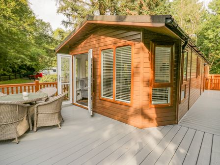 A wooden cabin with glass doors open to a deck with wicker chairs and a glass table surrounded by trees at Willows Lodge White Cross Bay near Windermere