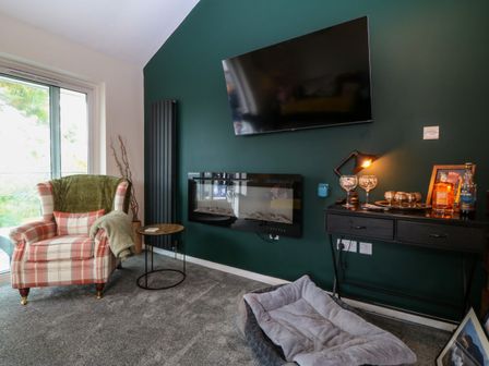 A living room with a plaid armchair near a sliding glass door and a green wall with a mounted TV and electric fireplace at The Sail Loft in Y Felinheli