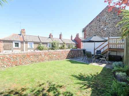 An outdoor garden area with green grass a stone wall and a patio set with table umbrella and chairs at Crab Corner in Wells-Next-The-Sea