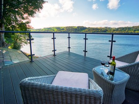 A balcony with two wicker chairs a small glass table with a bottle of wine and two glasses overlooking a lake and hills at Arenig - Bala Lake in Bala