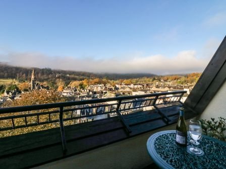 A balcony with a metal table bottle and glass overlooking a town with a church and hills at Loughrigg in Ambleside
