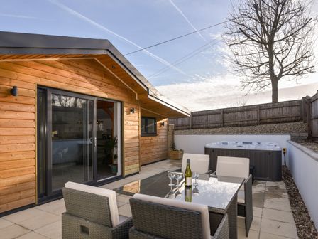 A patio area with a glass table and four chairs a bottle and glasses on the table a wooden cabin and a hot tub in the corner at The Stables @ Merrydown in Axminster