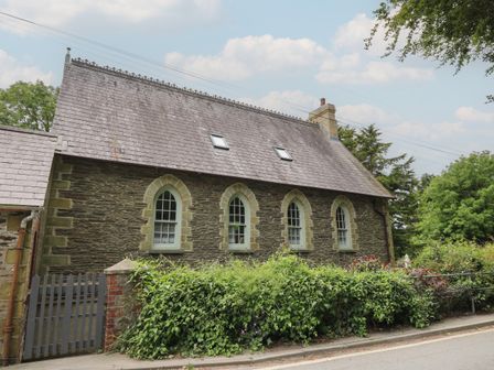 A stone house with arched windows and a slate roof with skylights behind greenery and a road at Upper Vestry in Llangrannog