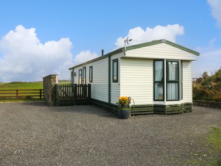 A single white prefabricated house with green trim on a gravel area near a green wooden fence and grassy fields at Cae Carreg in Cemaes Bay