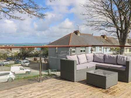 An outdoor balcony with a patio sofa set and glass railing overlooking houses and the sea at The Mordon in Newquay