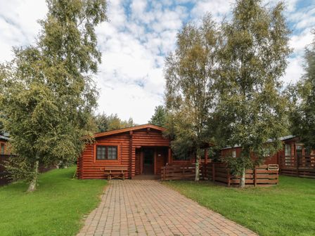 A log cabin with a paved pathway and trees in front at Welcome Hjem Log Cabin in Morpeth near Felton
