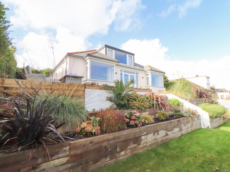 A exterior view of a house with multiple windows and a landscaped garden with a variety of plants and a wooden retaining wall at Higher Bolenna in Perranporth