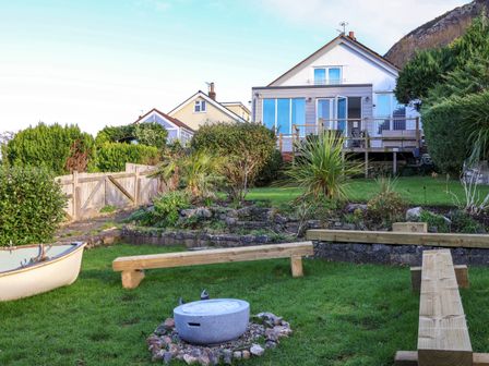 A backyard with a grass lawn a small boat a fire pit wooden benches plants shrubs and a house with a deck at Awel Y Mor in Prestatyn