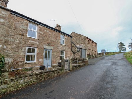 Stone cottages beside a narrow paved road with a metal gate and planters at Lime Tree Cottage in Kirkby Stephen