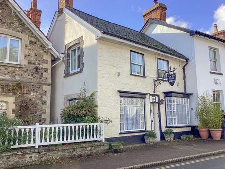 An old stone building with a sign reading The Old Lace Shop and white windows next to a white building with potted plants at The Old Lace Shop in Beer