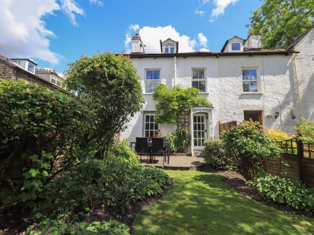 Rear view of a white house with a garden patio and outdoor table with chairs surrounded by shrubs at Brook Cottage in Windermere
