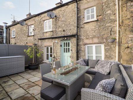 An outdoor patio with a wicker sofa and table set beside a stone building with windows and a mint green door at Market Square Maisonette in Kirkby Lonsdale