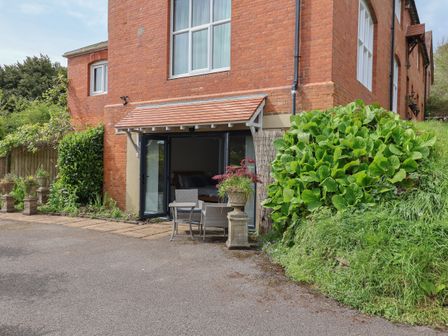Red brick building with open patio doors leading to a room with a bed outdoor chairs and a table beside green plants at The Hideaway in Uplyme near Lyme Regis
