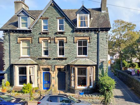 A stone house with two front doors and multiple windows on a street with parked cars at Cranford House in Keswick