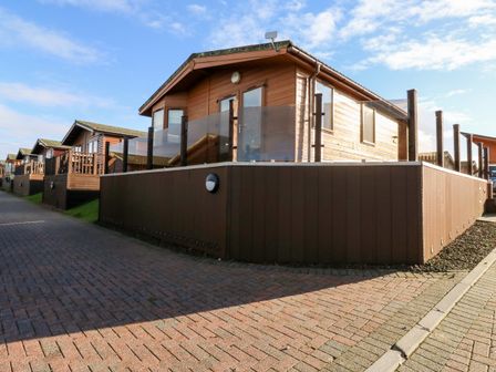 A row of wooden cabins with glass balconies along a paved walkway at Serenity Sunset in Ilfracombe
