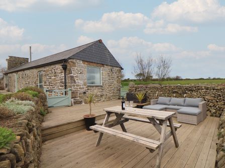 An outdoor patio with a wooden picnic table, a gray cushioned sofa, potted plants, and a stone building in the background at Long Barn in Castle Gate near Longrock