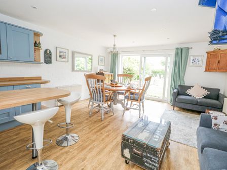 A dining area with round wooden table and chairs next to sliding glass doors leading outside with sofas and kitchen stools in Mermaid Cottage in Wakeham Portland