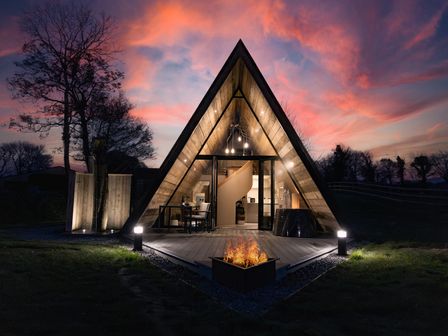 An illuminated triangular cabin with a fire pit in front and trees in the background at The Tri-Pod in Pantcefn near Llanarth