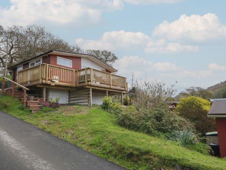 A raised red cabin with a wooden deck on a grassy slope near a road at Y Goedlan in Tywyn
