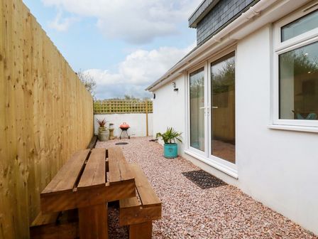 A patio area with a wooden picnic table a small barbecue grill and potted plants outside a house at Beagle Cottage in Fraddon