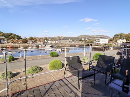 A balcony with three chairs and a bottled drink overlooking a marina with boats and buildings at Sea Breeze in Porthmadog