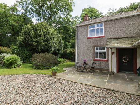 A house exterior with a pebble driveway a stone patio with a small table and two chairs and trees in the background at Gardeners Cottage in Millom Cumbria
