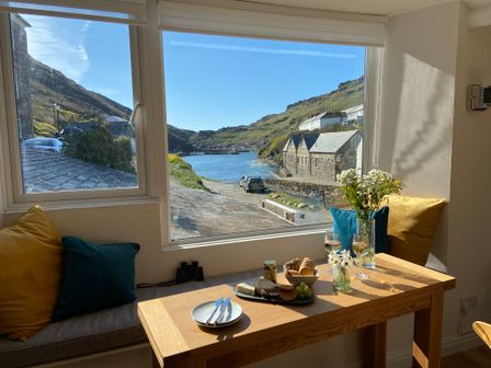 A dining table set with food, drinks, and flowers by a window with a view of a coastal village and hills at Harbour Light in Boscastle