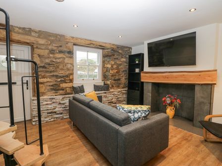 A living room with a gray sofa a wall-mounted TV exposed stone walls and a decorative fireplace at Cosy Logs Cottage in Cark In Cartmel