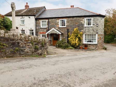 A stone building with white framed windows a wooden door and plants outside by a road at Mill Cottage in St Neot