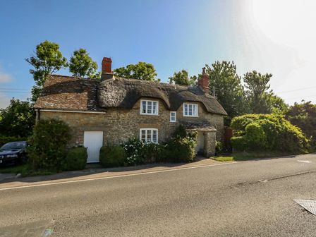 A stone cottage with a thatched roof beside a road with trees and bushes in Burton Bradstock
