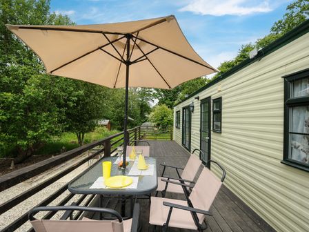 A wooden deck with a glass table and beige chairs under a large beige umbrella beside a cream mobile home surrounded by trees at Little Malory in St Giles-On-The-Heath
