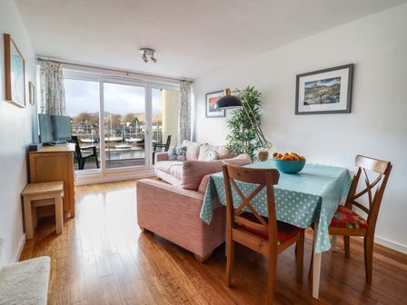 A dining area with a table covered by a blue tablecloth and wooden chairs next to a pink sofa and sliding glass doors overlooking boats at Branwen in Porthmadog