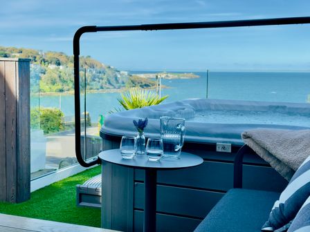 A patio with a hot tub a small round table with a jug and two glasses and a couch next to a glass railing overlooking the sea at Bayvista in Carbis Bay