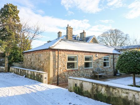 A stone cottage with snow on the roof and ground with a bench outside Wellington Lodge Cottage in Middleton Tyas