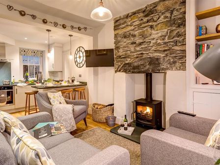 A living room with a stone fireplace a wood stove grey sofas and a kitchen island with stools at Cosy Cottage in Ambleside