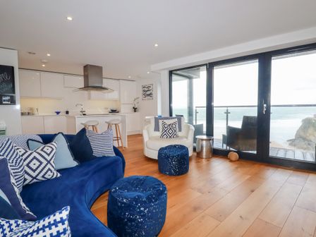 A living room with blue sofa white armchair kitchen and large windows overlooking the ocean at Towan Penthouse in Newquay