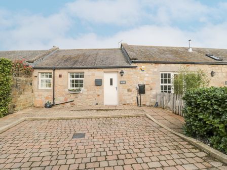 A stone cottage with a white door two windows and a paved driveway with shrubs at Little Tern in Seahouses