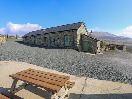 A stone building with multiple green doors and windows beside a gravel driveway with a wooden picnic table in the foreground at Ty Den in Llanberis