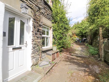 An exterior view of a stone house with a white door and window alongside a narrow garden path with outdoor seating at the end at Fell View in Ambleside