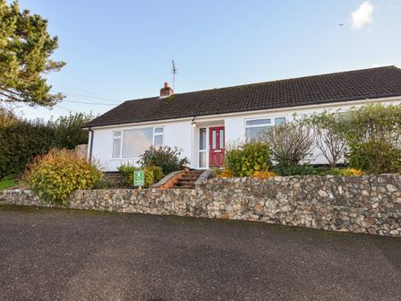 A single-story house with a red door and white walls surrounded by bushes and a stone wall at Hook Farm Cottage in Uplyme