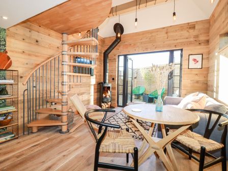 A living area with wooden spiral staircase a round table with chairs a stove and an open door leading to a patio at Little Beach Cabin in Crantock