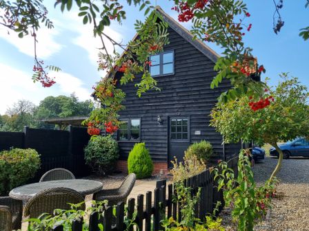 An outdoor seating area with a round table and chairs near a black wooden house with plants and a gravel driveway at The Hayloft in Haddiscoe