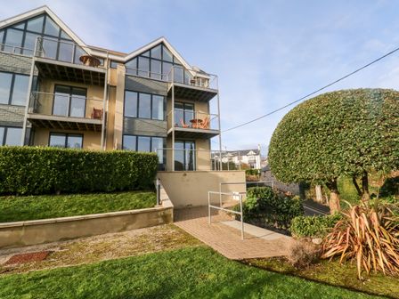 A modern three-story building with balconies and large windows a paved pathway leading to the entrance and trimmed bushes and trees at Ocean View in St Ives